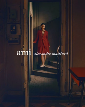 A fashionable woman in a red suit stands on a stairwell in an indoor setting with dim lighting for a fashion editorial shot.