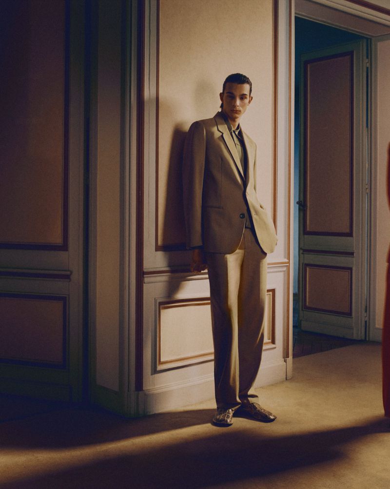 An adult man in a beige suit stands in a corner with decorative molding, his hand in his pocket in an indoor studio shot.