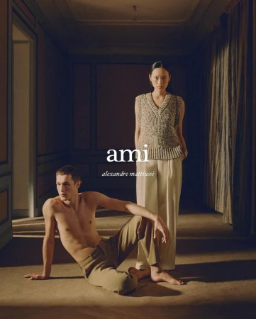 Two fashion models pose in an elegant, dimly lit interior hallway. One sits shirtless, the other stands in a knit sweater vest.