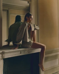 Model sitting barefoot on a marble fireplace mantel wearing a striped button-down shirt, reflecting in an ornate wall mirror.