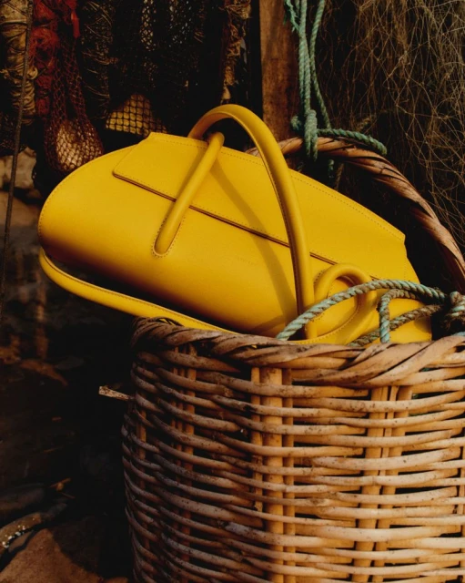 Eye-level shot of a yellow leather handbag inside a wicker basket, set against a background of fishing nets and wooden planks.