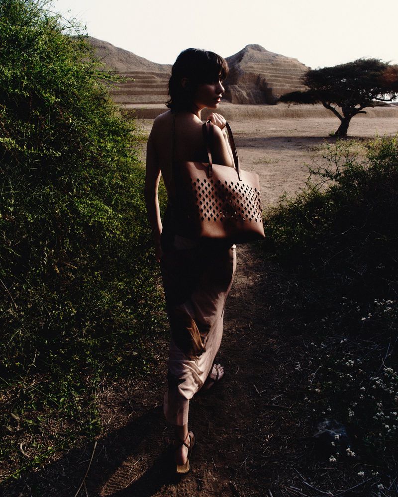Woman with a brown tote bag walking on a dirt path surrounded by green bushes with a hill and a tree in the background.