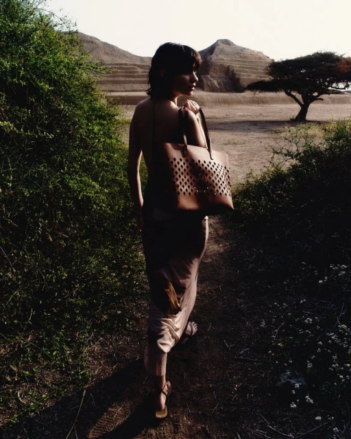 Woman with a brown tote bag walking on a dirt path surrounded by green bushes with a hill and a tree in the background.