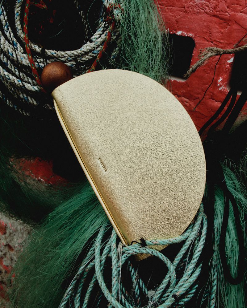 Close-up of a small, off-white leather purse with rope details, placed against a background of fishing nets and weathered red buoy.