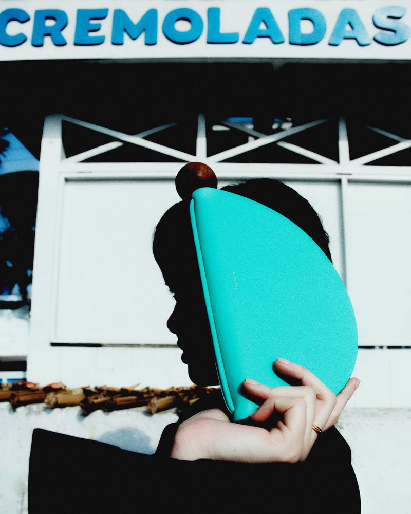 Side view of a person holding a teal clutch near an outdoor building with 'CREMOLADAS' text sign during daytime, shot in natural light.