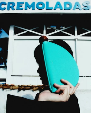 Close-up profile silhouette of a person holding a bright turquoise curved leather handbag in front of their face, outdoors against a white building