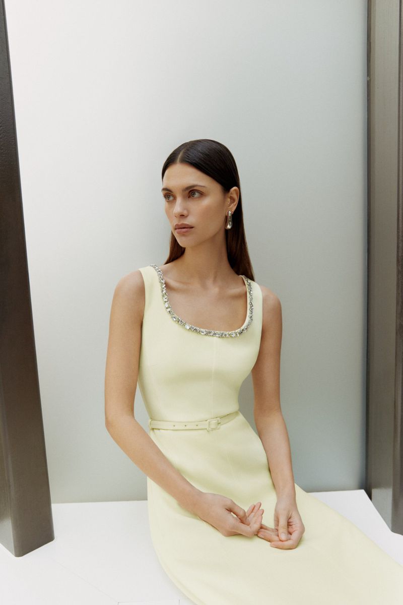 Brunette woman poses in a light yellow sleeveless dress with a jeweled neckline, seated against a white wall in a minimalist setting.