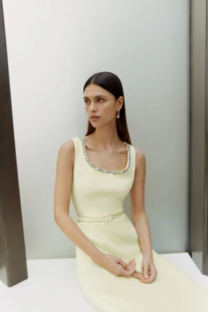Brunette woman poses in a light yellow sleeveless dress with a jeweled neckline, seated against a white wall in a minimalist setting.