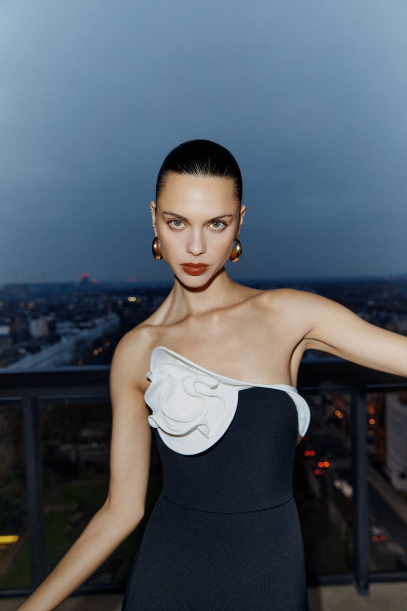 Woman in elegant black and white dress with modern gold hoop earrings poses on a rooftop at dusk, with cityscape in soft focus.