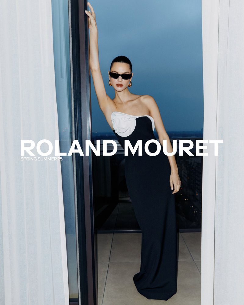 Elegant woman poses in black strapless dress with white floral detail for Roland Mouret Spring/Summer 23 campaign, in doorway setting.