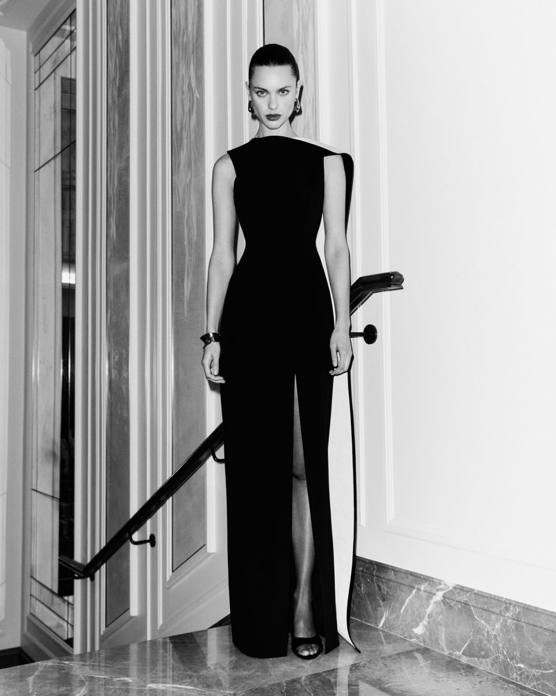 Elegant woman in a black floor-length dress with a thigh-high slit, standing indoors with marble floors and stairs, posing for a fashion shot.
