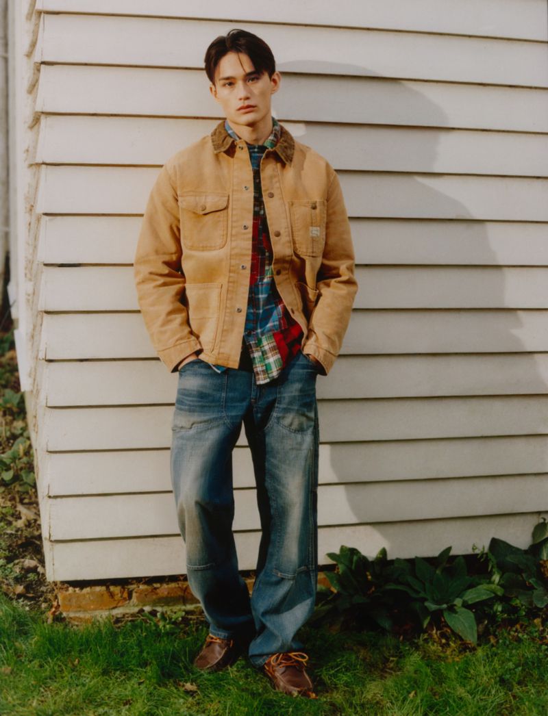 A young man poses outdoors, wearing a tan jacket, plaid shirt, and blue jeans, standing in front of a white clapboard building and green grass.