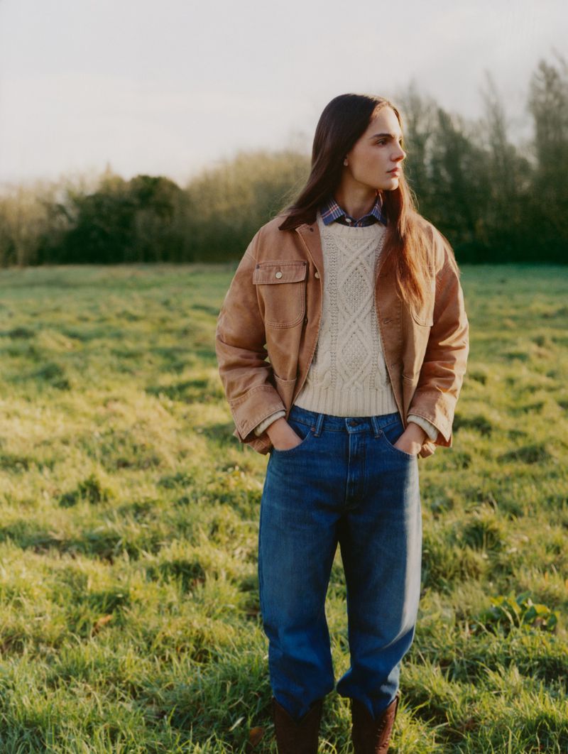 Woman in jacket and jeans stands in grassy field. Outdoor fashion shot with green grass, trees, and natural light. Warm daytime.