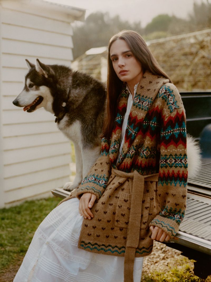 Young woman in patterned cardigan poses with a husky dog near the back of a pickup truck in an outdoor, rural setting.