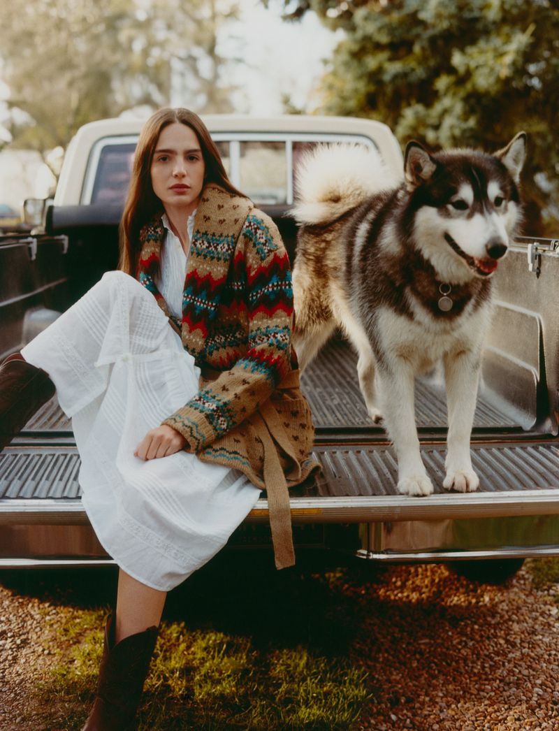 Young woman in white dress and patterned cardigan sits in a vintage truck bed with an Alaskan Malamute dog outdoors.
