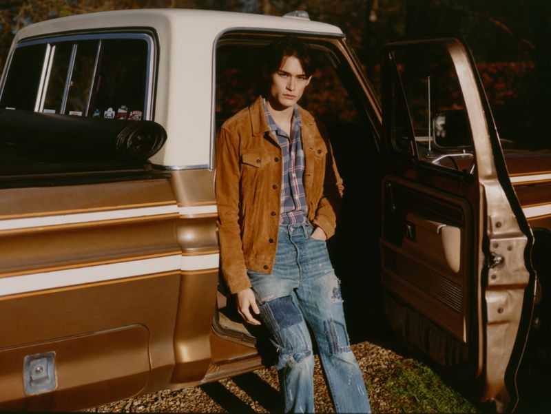 A young man in a brown corduroy jacket and patchwork jeans leans against a vintage pickup truck with a two-tone brown and white paint job.