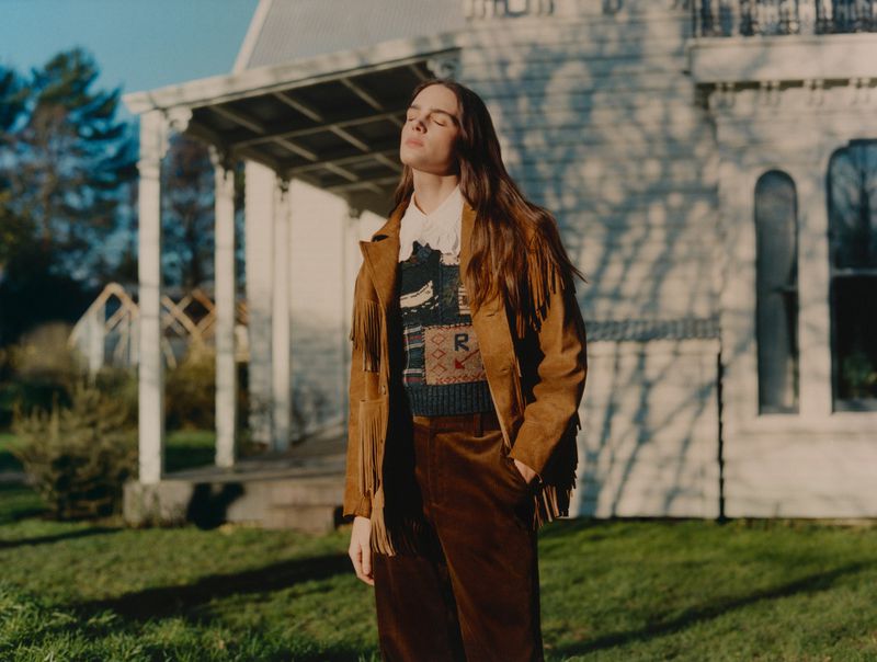 A woman with long brown hair stands in a grassy yard in front of a white house, wearing a tan fringed jacket and brown pants, basking in sunlight.