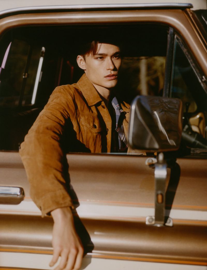 Male fashion model in a tan suede jacket sits inside of an early model pick-up truck. Natural light illuminates the model's features.