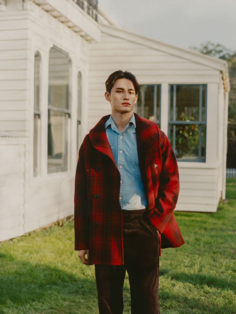 Portrait of a young man in a red plaid coat, light blue shirt, and brown pants, standing in front of a white house and green lawn.