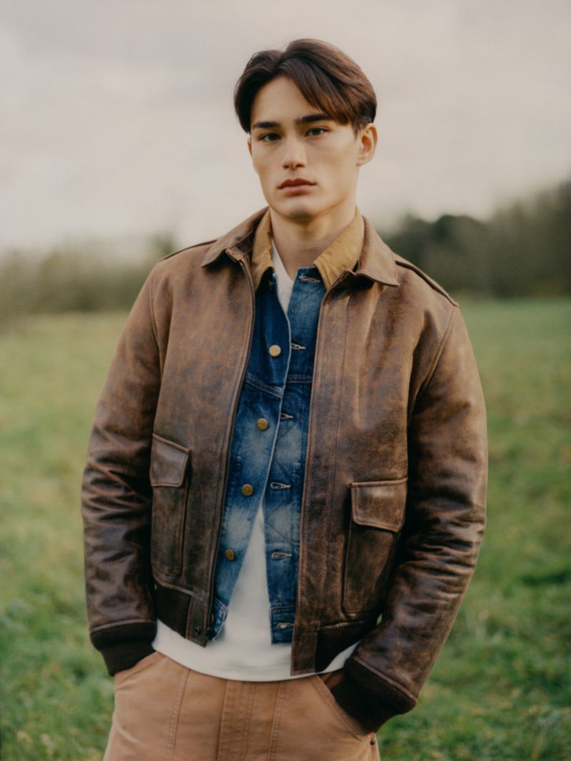 Portrait of an adult male with dark hair wearing a brown bomber jacket over a denim jacket outdoors in a grassy field.