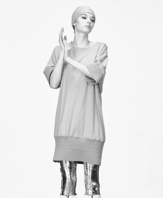 Black and white fashion shot of a young female model wearing a dress, matching head covering, and metallic boots in a studio setting.