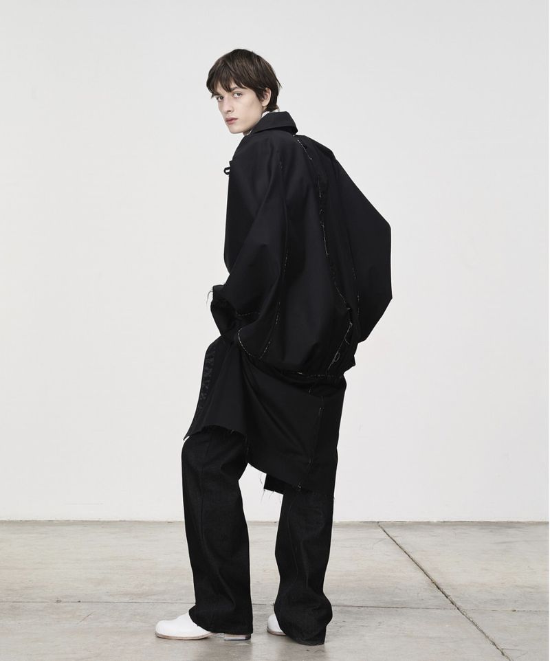 A slender man in a black overcoat and pants is photographed in a minimalist studio with a white wall and concrete floor. He glances back.