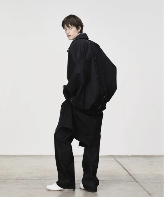 A slender man in a black overcoat and pants is photographed in a minimalist studio with a white wall and concrete floor. He glances back.