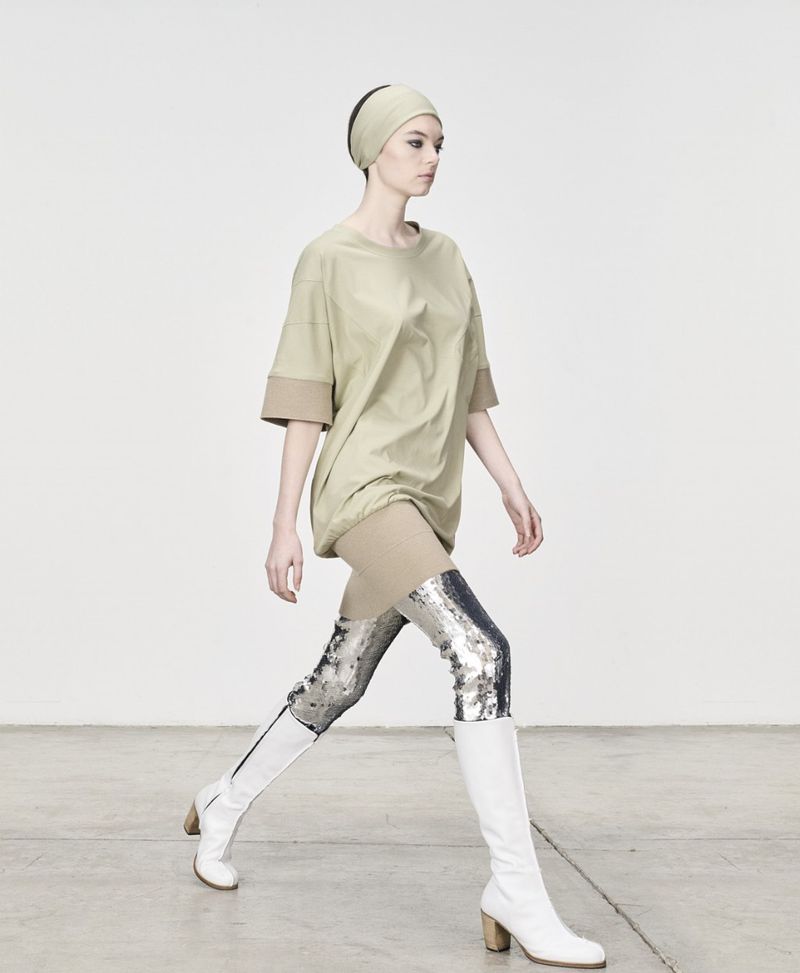 Fashion model strides confidently in an oversized khaki top, metallic leggings, and white boots against a clean white backdrop. 