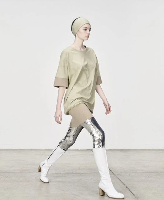 Fashion model strides confidently in an oversized khaki top, metallic leggings, and white boots against a clean white backdrop. 