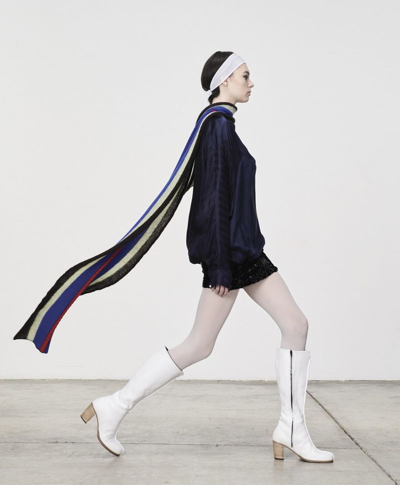 Fashion shot of a young adult woman in a blue top, black skirt, white boots, and a flowing scarf, posing against a bright studio backdrop.
