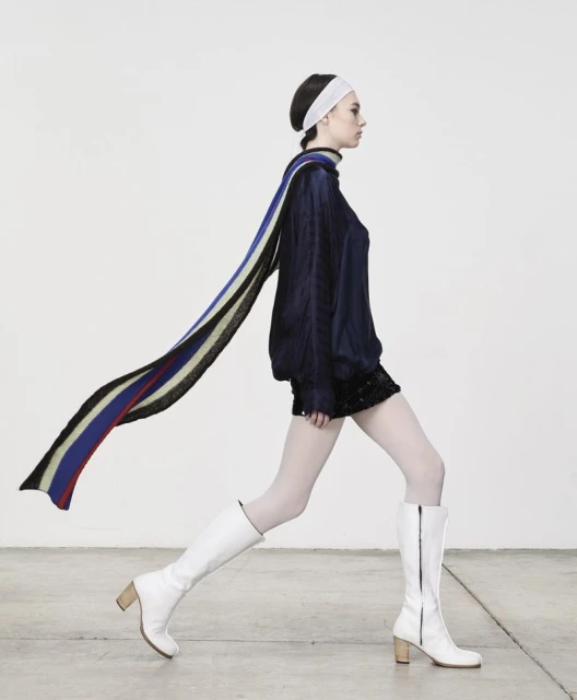Fashion shot of a young adult woman in a blue top, black skirt, white boots, and a flowing scarf, posing against a bright studio backdrop.