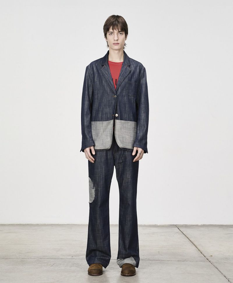 Full-length shot of a young adult man in a blue denim suit with patches, standing against a solid white background, and wearing brown shoes.