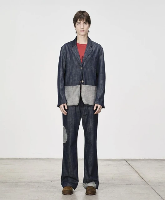 Full-length shot of a young adult man in a blue denim suit with patches, standing against a solid white background, and wearing brown shoes.