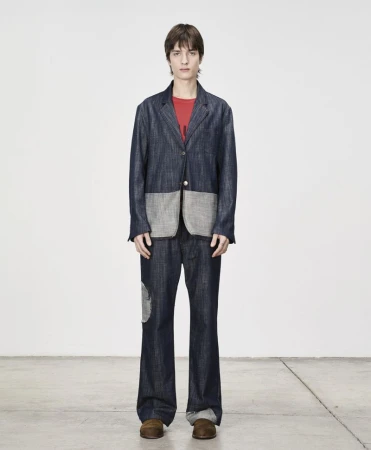 Full-length shot of a young adult man in a blue denim suit with patches, standing against a solid white background, and wearing brown shoes.