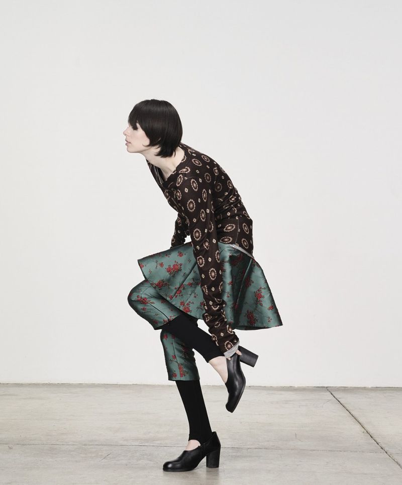 Woman in a patterned top and green skirt changing shoes, studio shot. Dark hair, bright white wall, and grey floor. Fashion editorial style.