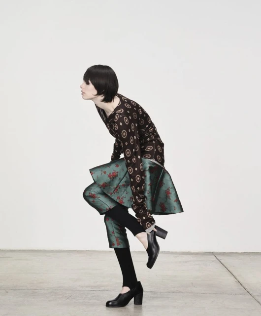 Woman in a patterned top and green skirt changing shoes, studio shot. Dark hair, bright white wall, and grey floor. Fashion editorial style.