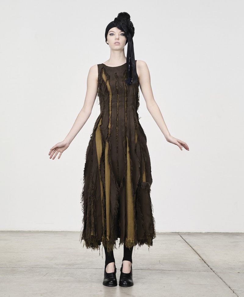 A solitary adult female model poses in a sleeveless, earth-toned textured dress against a bright studio backdrop, complemented by dark shoes.