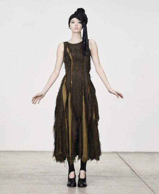A solitary adult female model poses in a sleeveless, earth-toned textured dress against a bright studio backdrop, complemented by dark shoes.