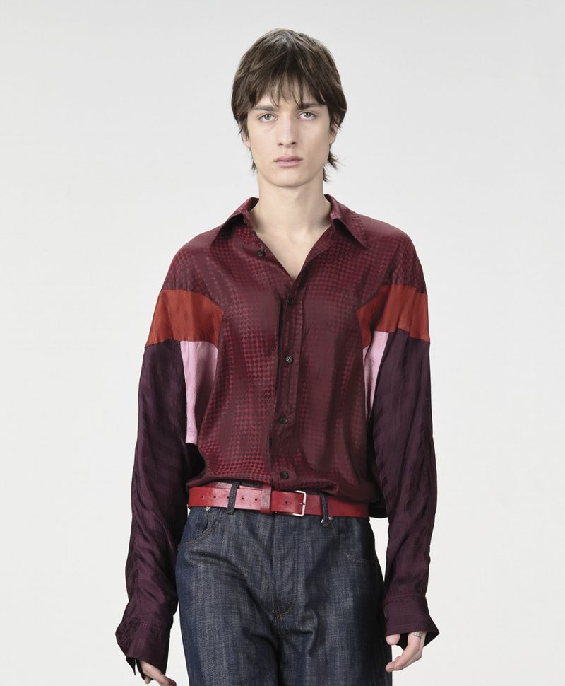 A slender male model poses in a patterned, color-blocked button-down shirt and jeans against a clean white backdrop in a studio shot.