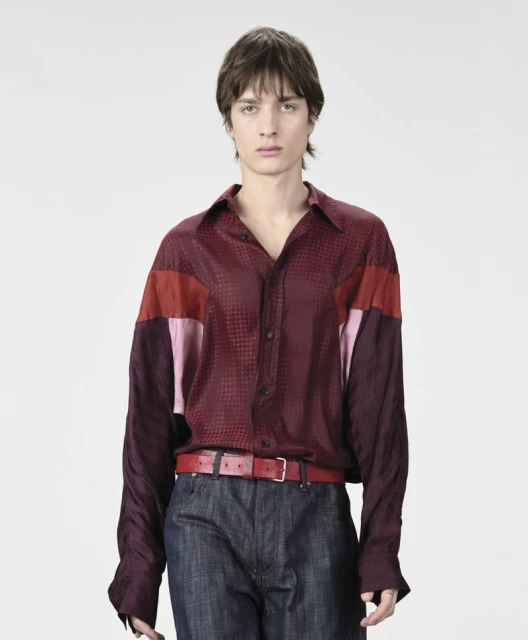 A slender male model poses in a patterned, color-blocked button-down shirt and jeans against a clean white backdrop in a studio shot.