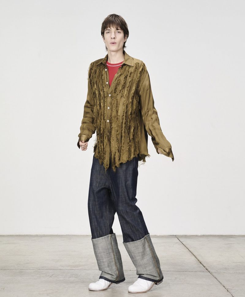 Full-shot studio portrait of a fair-skinned young adult wearing an open button-down shirt, jeans, and white shoes in a white studio.