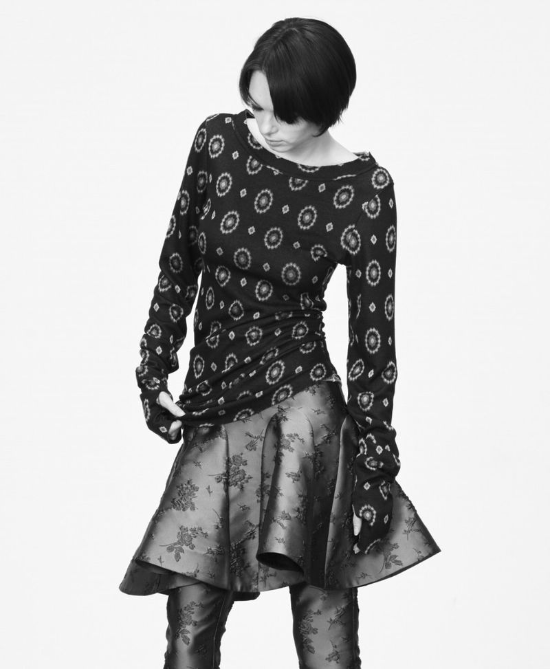 Monochrome fashion shot: a woman in a patterned long-sleeve top and flared skirt poses against a plain background, looking down.