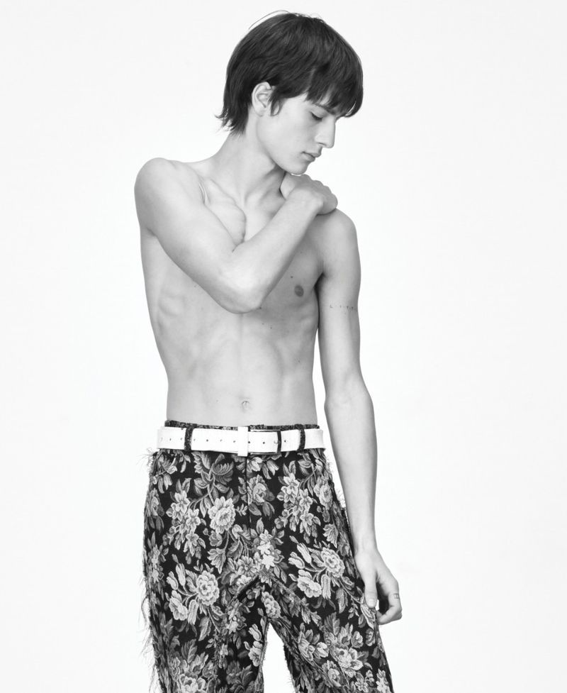 Monochrome studio shot of a shirtless young male fashion model wearing floral pants and a white belt against a white background.