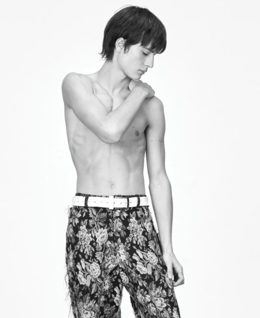 Monochrome studio shot of a shirtless young male fashion model wearing floral pants and a white belt against a white background.