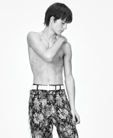 Monochrome studio shot of a shirtless young male fashion model wearing floral pants and a white belt against a white background.