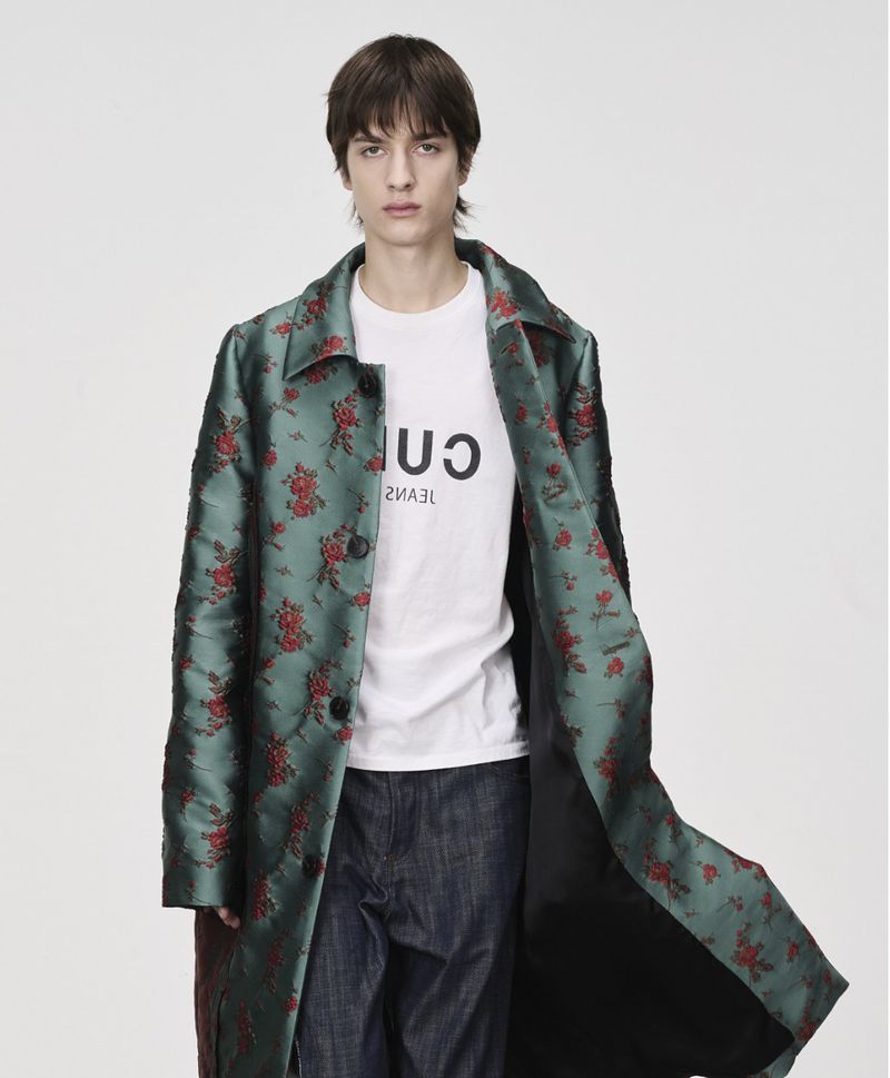 Man wearing a floral print trench coat and jeans against a white backdrop in a studio fashion shoot.