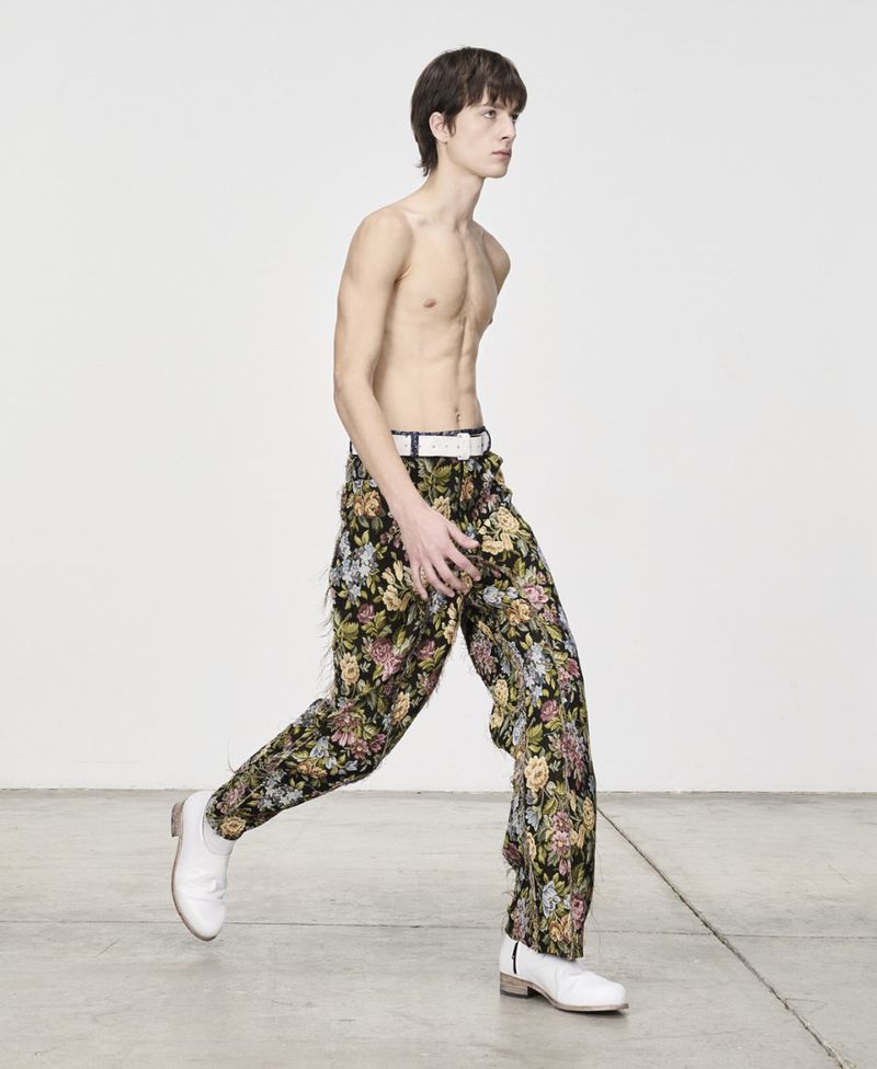 A slender, bare-chested adult male with dark hair is in motion, wearing floral trousers and white shoes, set against a bright white backdrop.