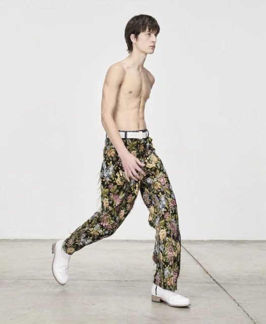 A slender, bare-chested adult male with dark hair is in motion, wearing floral trousers and white shoes, set against a bright white backdrop.