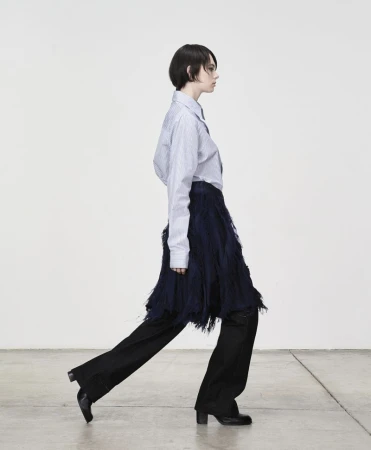 Model in profile wearing a striped button-down shirt and fringed skirt against a plain wall