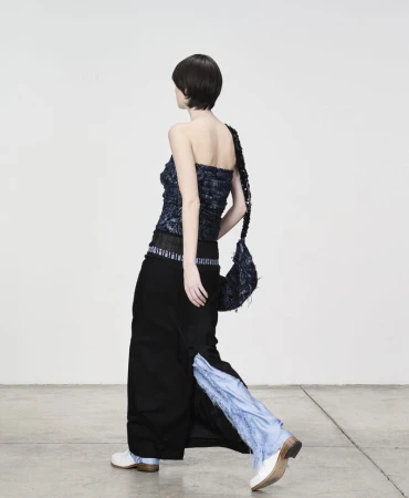 Model with dark hair walking away wearing a dark textured top, long black skirt, and white shoes against a plain wall.